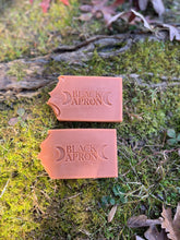 Load image into Gallery viewer, Two peach-orange colored bars of soap with 'Black Apron' branded soaps on a mossy background
